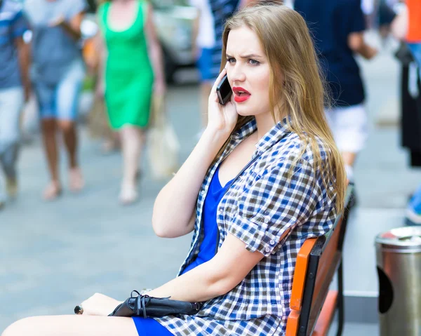 Технологии и коммуникации. Молодая блондинка красивая девушка спорит и разговаривает по мобильному телефону. Крупный план портрета улыбающейся студентки с красными губами разговаривающей по телефону на фоне улицы . — стоковое фото