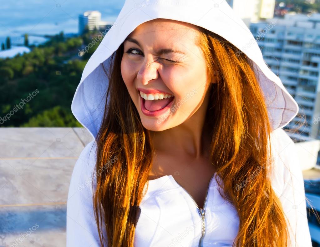 Close up sunset fashion portrait of young teenage girl and sitting on the roof with amazing sea view. Sensual cute fresh happy face, long colored hair, crazy positive emotions. Wearing white hoodie.