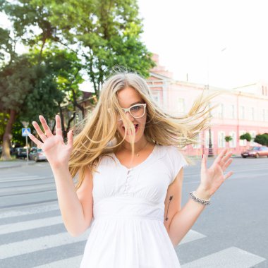 Close up portrait of exciting happy blondy young woman that is dancing in the street, wearing white dress and clear glasses. Crazy emotions, positive lifestyle. Amazing summer day.