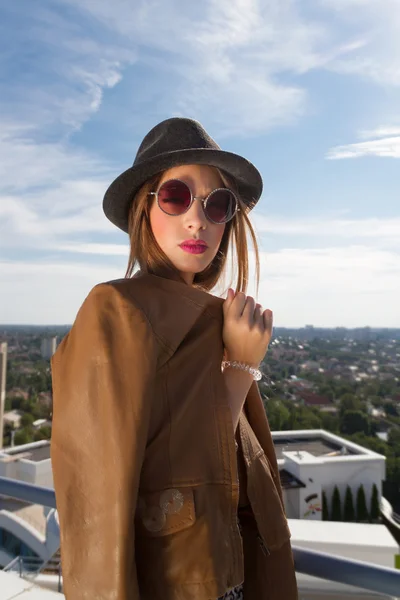 Adolescente urbana con sombrero posando en una chaqueta de cuero, en la parte superior de un edificio.Retrato de moda al aire libre de la señora, con sombrero vintage, gafas de sol y chaqueta de cuero, increíble vista de la ciudad desde el techo . — Foto de Stock