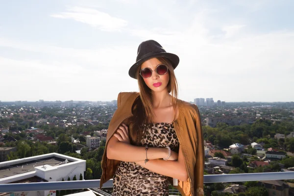 Adolescente urbaine avec chapeau posant dans une veste en cuir, sur le dessus d'un bâtiment.Portrait de mode extérieur de dame, portant un chapeau vintage, des lunettes de soleil et une veste en cuir, vue imprenable sur la ville depuis le toit . — Photo