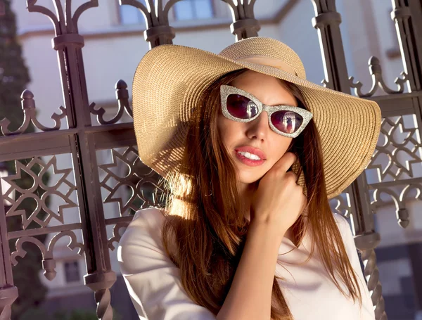 Portrait of pretty cheerful woman wearing white dress and straw hat in sunny weather day. Walking at summer park, smiling by beautiful fence in old city.Wearing cute pink dress,wide hat,cat glasses. — Stock Photo, Image