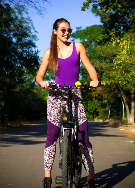 Сексуальная молодая женщина катается на велосипеде в городском парке. Happy girl on bike cycling outdoors in summer smiling of joy during outdoor activity.Wearing sexy sportswear, purple swimwear and trendy leggings — стоковое фото