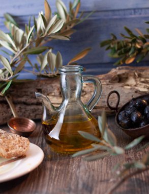 Görüntünün ortasında bir sürahi yağ, zeytin dalları, siyah zeytin ve dikey siyah yağlı tam buğday ekmeği olan kompozisyon.