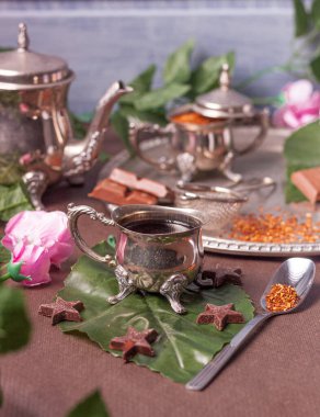 Çiçeklerle süslenmiş bir masa, çaydanlık ve çay için bir sürahi gümüş çay, çikolata ve tepside de çay..