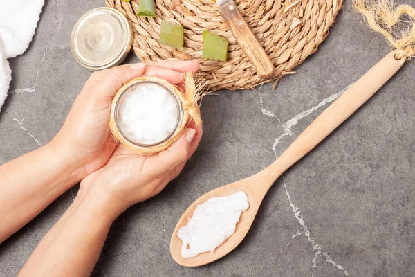 View of hands picking up a jar of aloe vera hand cream with a trivet with sliced aloe vera pieces and a spoon with hand cream