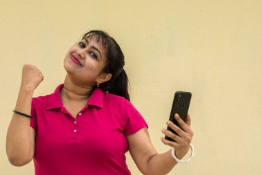 Hintli kadın model mutlu bir şekilde cep telefonuna bakıyor sarı arka planda gülümseyen hafif bir yüz ve metin için kopya alanı var. Mutlu ruh hali konsepti.