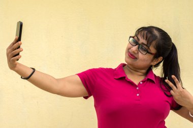 Cep telefonuna bakan Hintli kadın model mutlu bir şekilde selfie çekiyor sarı arka planda gülümseyen bir yüz ve metin için fotokopi alanı. Mutlu ruh hali konsepti.