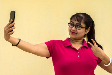 Hintli kadın model mutlu görünüyor. Hafif gülümseyen yüzüyle. Sarı arka planda akıllı telefonlu selfie çekiyor.