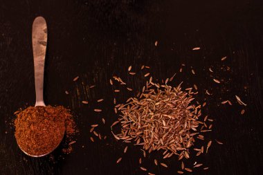 Bir çay kaşığı dolusu jeera ya da kimyon tozu ve siyah tahta bir masanın üzerinde biraz çiğ kimyon tohumu. Seçici odak.