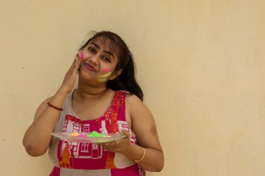 Holi renkleriyle dolu bir tabak taşıyan ve yüzüne renk uygulayan güzel bir Hintli kadın. Sarı arkaplan ile seçmeli odak.