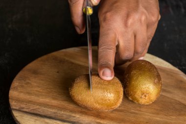 Bir erkek eli, siyah bir tahta yüzeyde iki olgun kivi meyvesini bıçakla kesecek. Seçici odak.