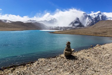 Gurudongmar Gölü 'nün zemininde kayalar ve arkasında mavi gökyüzü olan bir dağ manzarası. Gurudongmar Gölü, Hindistan ve dünyanın en yüksek göllerinden biridir. Hindistan 'ın Sikkim eyaletinde 17.800 feet yükseklikte yer almaktadır..
