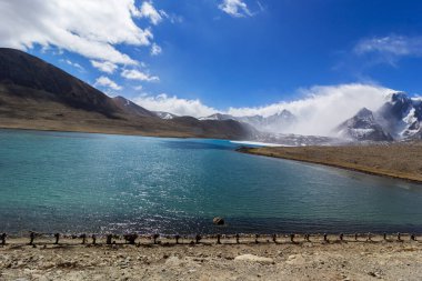 Gurudongmar Gölü 'nün zemininde kayalar ve arkasında mavi gökyüzü olan bir dağ manzarası. Gurudongmar Gölü, Hindistan ve dünyanın en yüksek göllerinden biridir. Hindistan 'ın Sikkim eyaletinde 17.800 feet yükseklikte yer almaktadır..