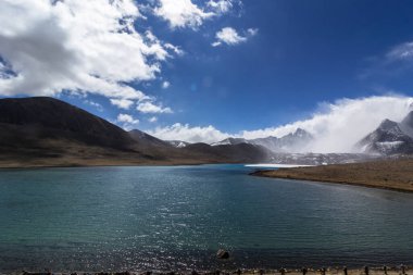 Gurudongmar Gölü 'nün zemininde kayalar ve arkasında mavi gökyüzü olan bir dağ manzarası. Gurudongmar Gölü, Hindistan ve dünyanın en yüksek göllerinden biridir. Hindistan 'ın Sikkim eyaletinde 17.800 feet yükseklikte yer almaktadır..