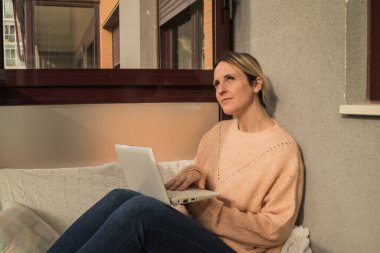 Terasta kanepede uzanmış, gülümseyen ve laptopta bilgi arayan bir kadın.