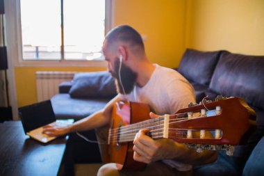 Ciddi sakallı bir adam kulaklıkla gitar çalmayı öğreniyor ve bir dizüstü bilgisayara danışıyor.