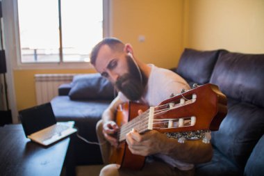 Sakallı konsantre bir adam. Kulaklıkla gitar çalmayı öğreniyor ve dizüstü bilgisayara danışıyor.