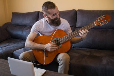Gülümseyen sakallı adam gitar çalmayı öğreniyor. Kulaklık ve dizüstü bilgisayarla.