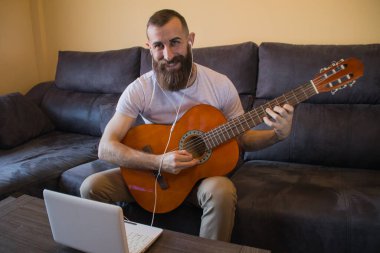 Gülümseyen sakallı adam gitar çalmayı öğreniyor. Kulaklık ve dizüstü bilgisayarla.