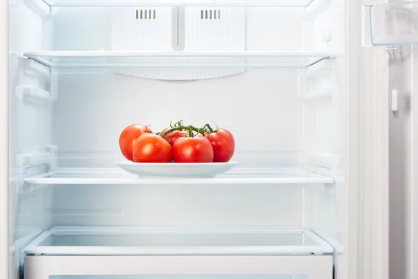 Zweig roter Tomaten auf weißem Teller im offenen leeren Kühlschrank — Stockfoto