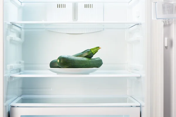 Два зеленых цуккини на белой тарелке в открытом пустом холодильнике — стоковое фото