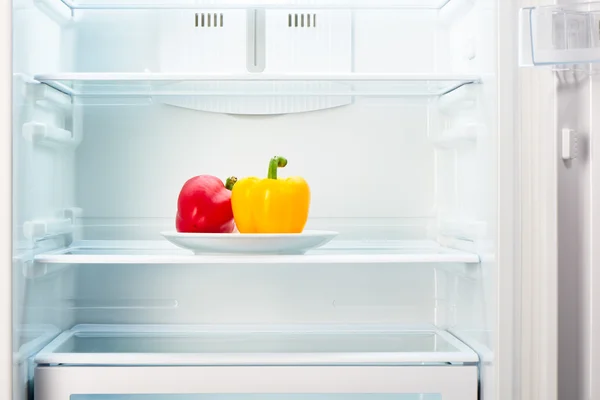 Red and yellow peppers on white plate in open empty refrigerator — Stock Photo, Image