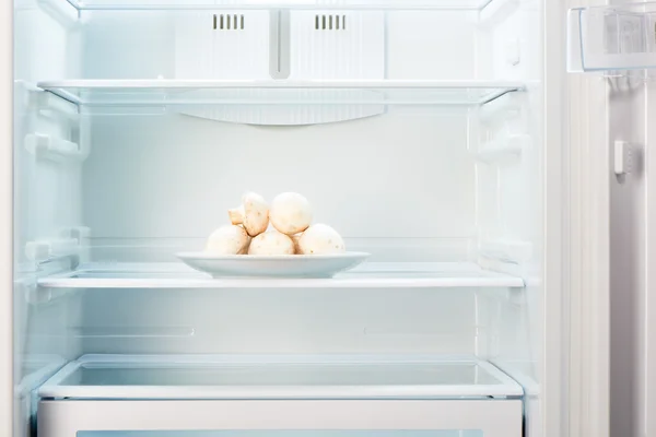 Fresh champignon mushrooms on white plate in open empty refrigerator — Stock Photo, Image