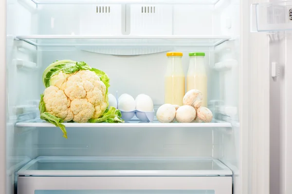 Cauliflower, white eggs, champignon mushrooms and two glass bottles of yoghurt on shelf of open empty refrigerator — Stock Photo, Image