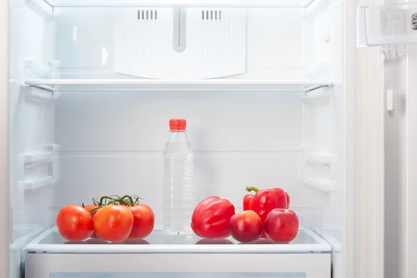 Zweig roter Tomaten, zwei rote Paprika, zwei zweifarbige orange und rote Pfirsiche und eine Flasche Wasser im Regal des offenen leeren Kühlschranks — Stockfoto