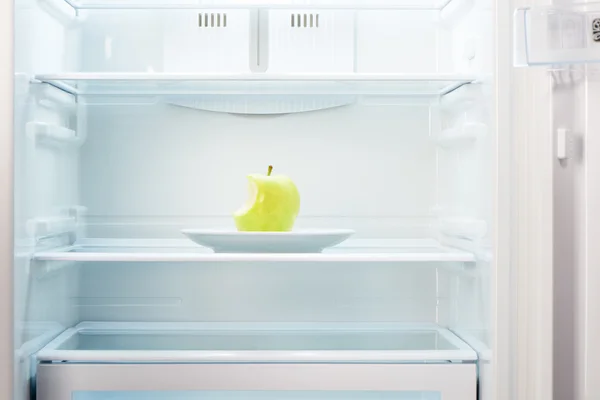 Green bitten apple on white plate in open empty refrigerator — Stock Photo, Image
