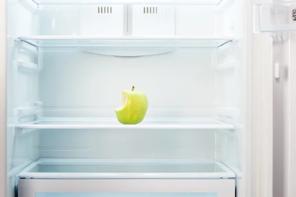 Green bitten apple on shelf of open empty refrigerator