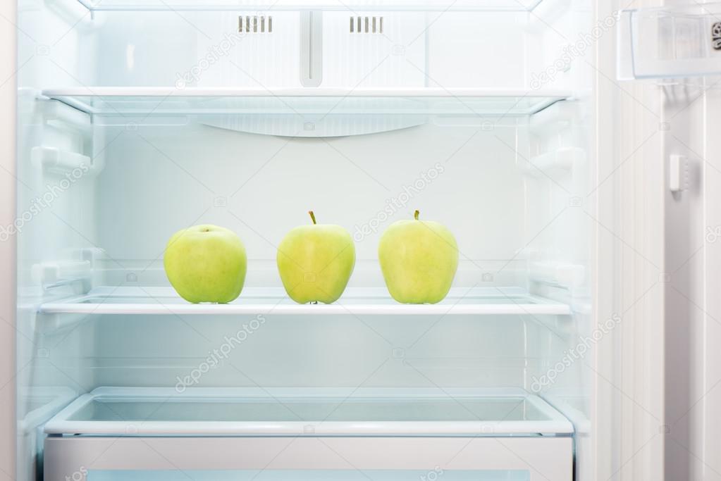 Three green apples on shelf of open empty refrigerator
