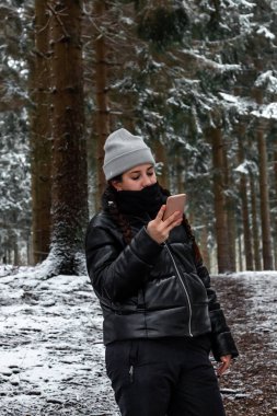 Danimarka 'da karlı bir ormanda akıllı telefon kullanan genç bir kadın sıcak kıyafetler giyiyor.