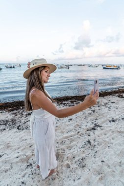 Şapkalı genç bir kadın sahilde selfie çekiyor.