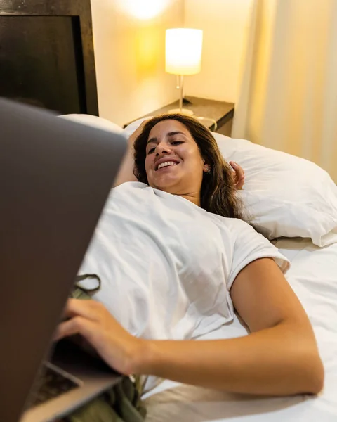 young hispanic woman lying in bed smiling using her laptop on her legs