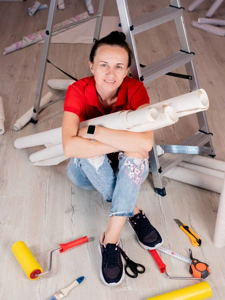 A woman is sitting on the floor in a room with rolls of Wallpaper. Concept of renovation in the apartment.