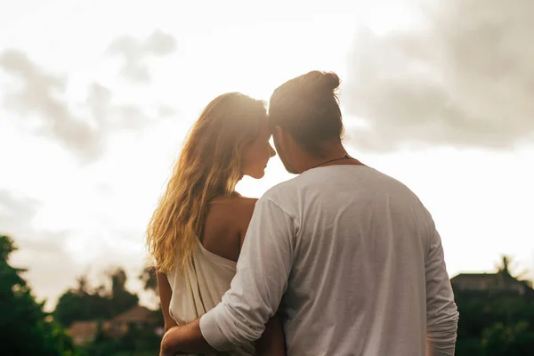 Uzun saçlı adam gün batımında narin kıvırcık kadınına sarılıyor.