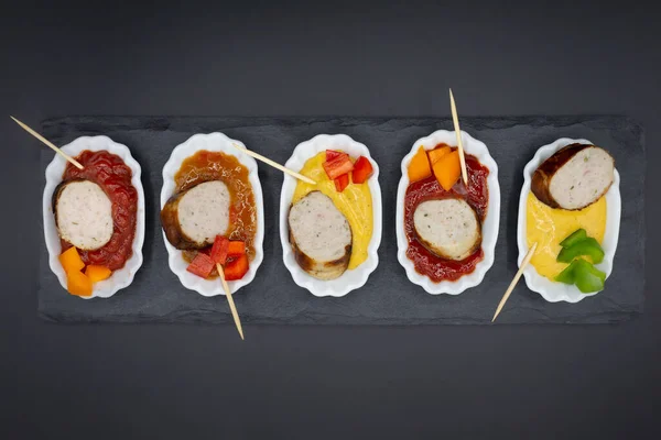 small plates with bratwurst and different sausages plus various dips. Arranged in a row with black background