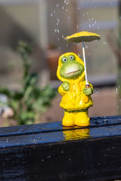 frog with yellow rain jacket stands in spring in the rain and is protected by a yellow rain umbrella, symbolizing unusual weather in season