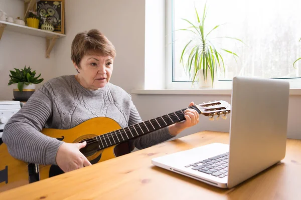 An elderly woman takes guitar lessons online. Senior retired woman studying online, watching online music lessons at home on a laptop. Online education.