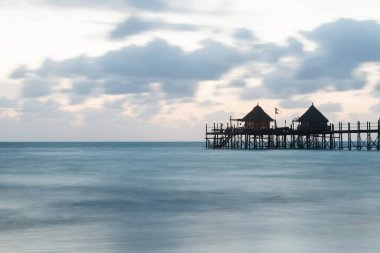 Güneş doğarken, Zanzibar adasında, tropikal bir sahilde ahşap iskele ve sazdan çatılar.