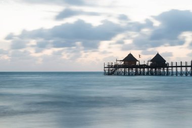 Wooden pier and thatched roofs on a tropical beach at sunrise, Zanzibar island clipart