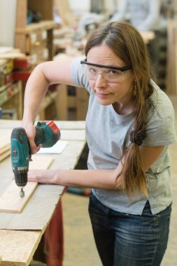 Matkaplı genç bir kadın atölyede ahşap bir kalasa delik açıyor.