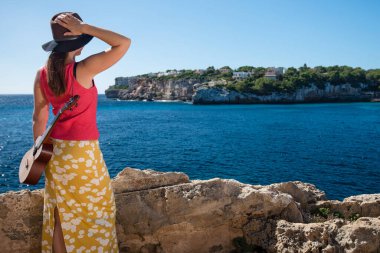 Omzunda gitar olan genç bir kadın sahilde durup deniz kenarında tatilinin tadını çıkarıyor.