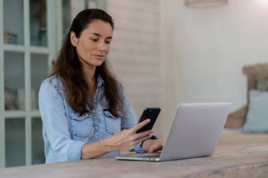 Masada oturan telefon ve bilgisayarla evde çalışan bir kadın. Evin arka planı bulanık. Ellerini kapat.