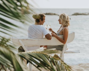 Mutlu çift sahilde şarap içer. Günbatımı arka planında aşık bir çift. Sevgililer Günü tatilde. Seyahat, aşk, gençlik yaşam tarzı konsepti. Sevgililer gününde birlikte seyahat edeceğiz. Gülümseyin ve birlikte şarap için. Palmiye yapraklarından vurulmuş.. 