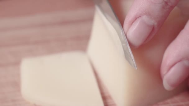 Le fromage de chèvre à pâte dure est coupé en fines tranches sur une planche à découper en bois. Cuisiner le petit déjeuner européen. Les mains féminines coupent le fromage avec un couteau. Concentration sélective. — Video