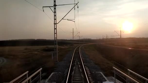 Vue depuis la fenêtre du dernier wagon du train jusqu'au coucher du soleil, les rails allant au loin, le virage et le pont ferroviaire traversant la rivière. Concept voyage en train. — Video