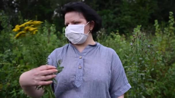 Kvinna i medicinsk mask utomhus. En person sniffar på en medicinblomma. Statisk kamera. Begreppet coronavirus. — Stockvideo